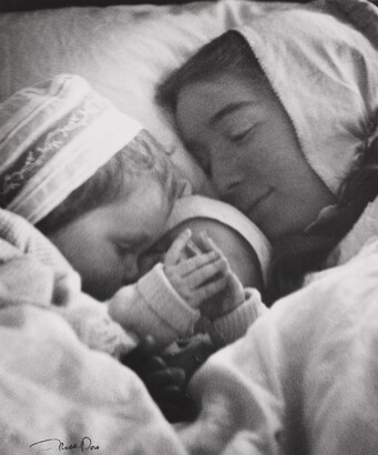 A black-and-white photograph of a White woman in a nightcap cuddling in bed with a toddler and an infant.