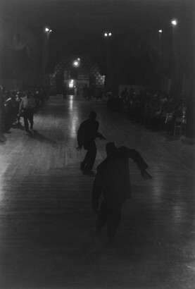 A dark black-and-white photograph of two male dancers in silhouette at the center of a dance floor.