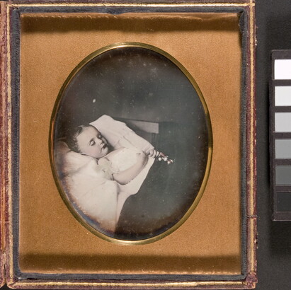 A black-and-white photograph in a gold frame of a child with closed eyes laying in bed with a toy in her clasped hands.