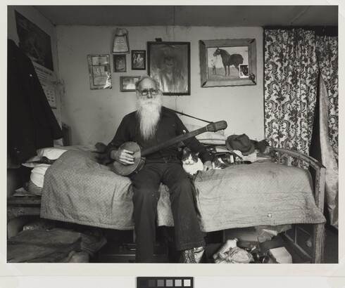 A black-and-white photograph of an elderly White man with a long white beard sitting on a bed in a cozy bedroom holding a banjo with one hand, the other hand around a cat lying next to him.