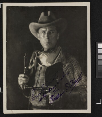 A black-and-white photograph of a White man in western attire holding a pistol in each hand, an autograph in blue in across the bottom.