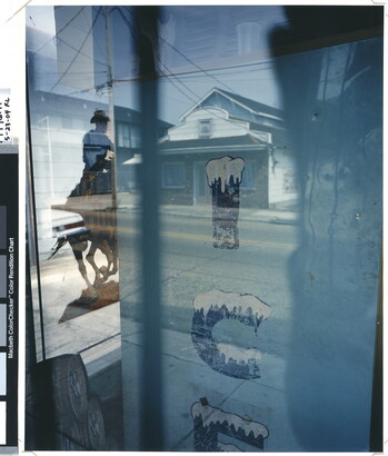 A color photograph taken through a window by the side of an ice machine and a poster of a man on a horse, as well as reflections of a house and other buildings.