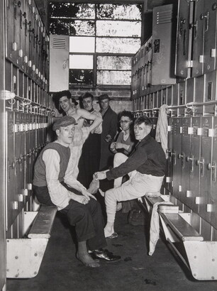 A black-and-white photograph of several men in various states of dress in the row of a locker room.