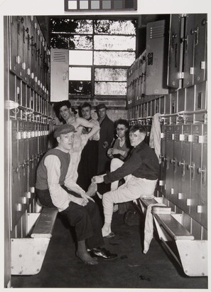 A black-and-white photograph of several men in various states of dress in the row of a locker room.