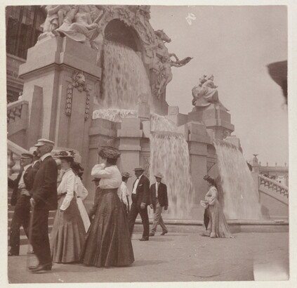 A sepia-toned photograph of women in long skirts and men in hats and jackets strolling by an elaborately sculpted facade with waterfalls.