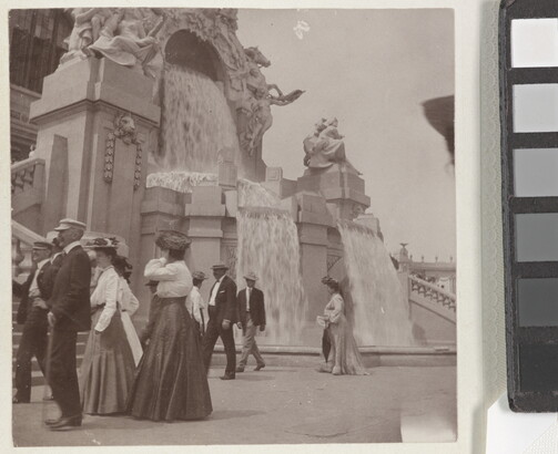 A sepia-toned photograph of women in long skirts and men in hats and jackets strolling by an elaborately sculpted facade with waterfalls.