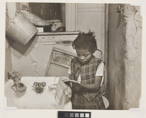 A black-and-white photograph of a young Black girl wearing a plaid vest and skirt washing her hands with a cloth in front of a bathtub.