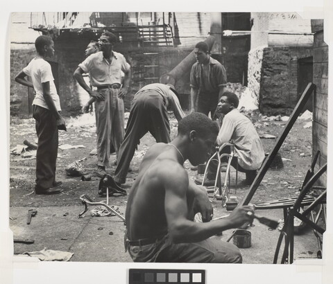 A black-and-white photograph of group of Black men talking and working in a rundown space littered with debris.