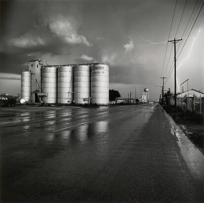 A black-and-white photograph of four grain elevators to the left of a wet road, a water tower in the far distance, and a flash of lighting behind power lines on the right.
