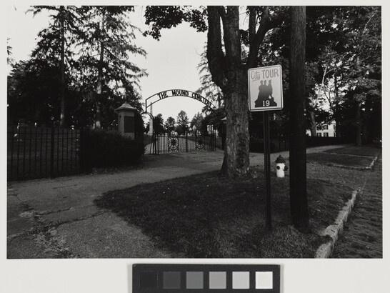 A black-and-white photograph taken from the street of a wrought iron gate a the entrance to a cemetery.
