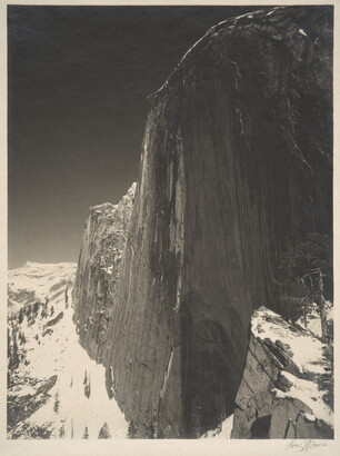 A black-and-white photograph of a steep cliff face and the snow-covered mountains around it.
