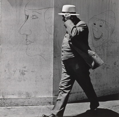 A black-and-white photograph of a man in a three-piece suit and hat walking past a wall covered with graffiti.