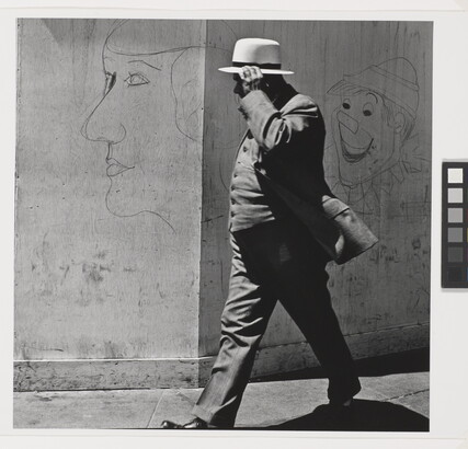 A black-and-white photograph of a man in a three-piece suit and hat walking past a wall covered with graffiti.