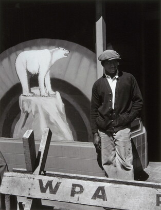 A black-and-white photograph of a Black man in a flat cap and dark cardigan behind a wooden barricade that says &quot;WPA&quot; and in front of a sign with a painting of a polar bear.