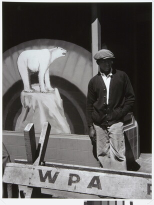 A black-and-white photograph of a Black man in a flat cap and dark cardigan behind a wooden barricade that says &quot;WPA&quot; and in front of a sign with a painting polar bear.