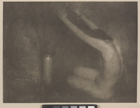 A soft-focus, black-and-white photograph of a nude White woman sitting on the floor next to a vase, her arms raised toward the flowers.