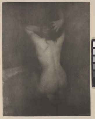 A dark, soft-focus, black-and-white photograph of a nude White woman kneeling with her back to the viewer and her hands in her hair.