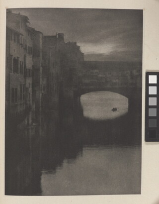 A sepia-toned photograph of a wall of tall buildings lining a river and an enclosed arched bridge stretching over the water.