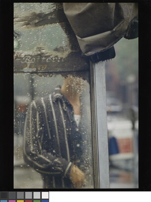 A color photograph of a man&#039;s torso through the rain-splatted glass of what looks like a bus shelter.