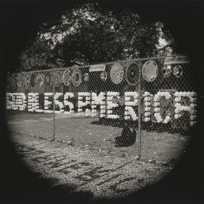 A black-and-white photograph of plastic cups arranged in a chain-link fence to spell out &quot;God Bless America.&quot;