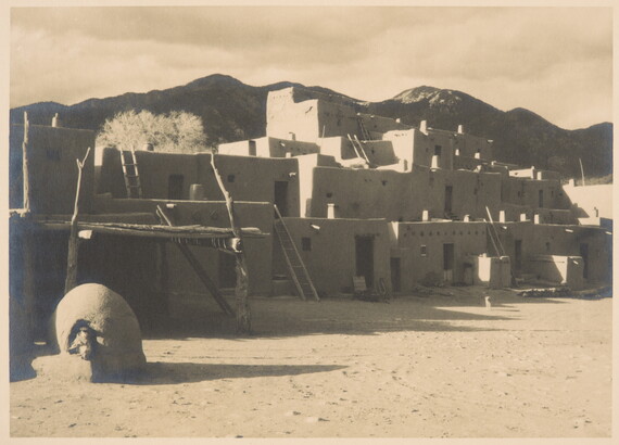 A black-and-white photo of a multi-level adobe pueblo with ladders leading from story to story and dark mountains in the background.