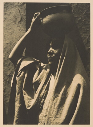 A black-and-white photograph of an Indigenous woman, face mostly in shadow, head and shoulders covered in a blanket or shawl, using her hand to balance a pot on her head.