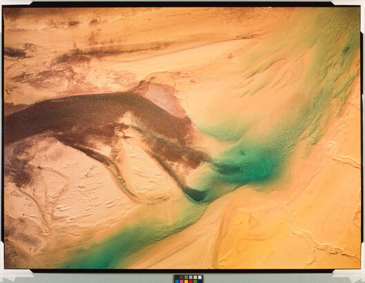A color photograph of sand dunes from the air with a bright blue-green streak across them.