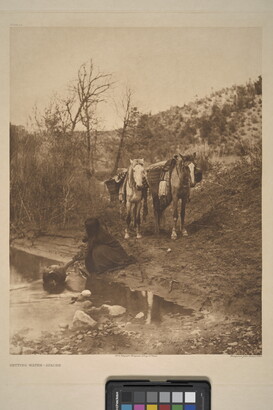 A sepia-toned photograph of an Indigenous person kneeling next to a stream to fill a jug with water as two packhorses wait in the background.