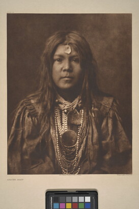 A sepia-toned portrait photograph of a young Indigenous person with long hair, a piece of jewelry on their forehead, multiple stings of beads around their neck, and wearing cultural dress.