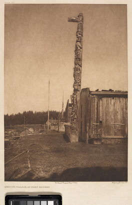 A sepia-toned photograph of a tall totem pole next to a wooden building with a forest in the background.