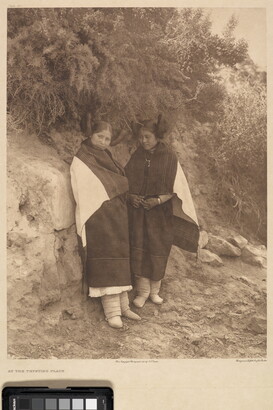 A sepia-toned photograph of two Indigenous women in traditional clothing leaning on a rock that has vegetation growing off the top of it.