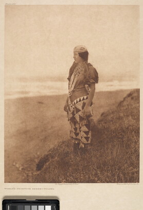 A sepia-toned photograph of a young Indigenous woman wearing traditional dress standing on a hill overlooking a plain.
