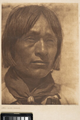 A close-up, sepia-toned portrait photograph of an Indigenous man with hair to his jawline, wearing a scarf around his neck held by a silver concho-style scarf slide.