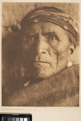 A sepia-toned portrait photograph of an older Indigenous person wearing a headband with a solid-colored cloth wrapped around their shoulders and under their face.