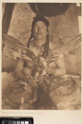 A sepia-toned photograph of a seated Native American man holding a large bird with its wings spread wide in one hand and a rattle in his other.