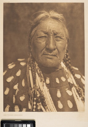A sepia-toned portrait photograph of an older Indigenous woman wearing a braids, elk-tooth shirt, and multiple necklaces.