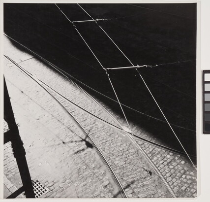 A black-and-white abstract photograph of tram tracks, a brick street, and wires divided diagonally by a hard shadow.