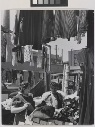 A black-and-white photograph of two light- to medium-skinned women smiling at each other in an open-air market, surrounded by clothing and boxes.
