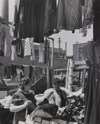 A black-and-white photograph of two light- to medium-skinned women smiling at each other in an open-air market, surrounded by clothing and boxes.