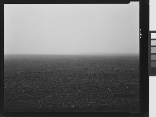 A black-and-white photograph of a dirt landscape with a hazy horizon and a subtle hill in the distance.