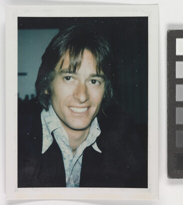 An informal color photograph of a smiling White man from the shoulders up with longish hair, wearing an open collar shirt and dark jacket.