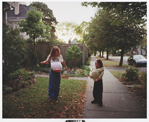 A color photograph of two young White girls playing in a front yard with balloons under their shirts mimicking pregnancy.