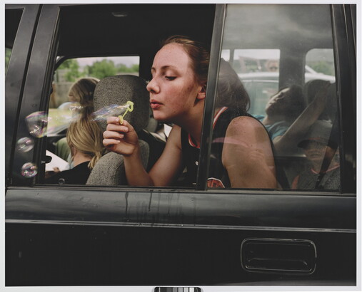 A color photograph of a White teenage girl blowing bubbles out of a backseat car window.