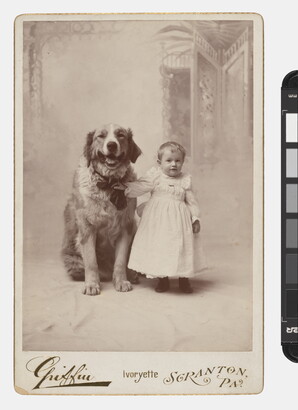 A black-and-white studio photograph of a toddler in a white dress standing next to a large dog and holding onto its collar.