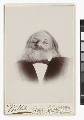 A black-and-white photograph of a deceased White man, from the shoulders up, with a full beard and eyes closed wearing a suit.