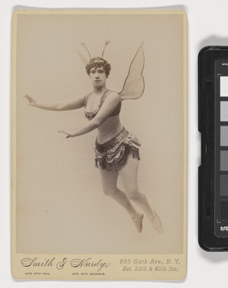 A sepia-toned studio photograph of a White woman dressed as a fairy, including wings and antennae, suspended in the air against a white background.