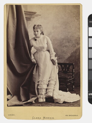 A sepia-toned studio portrait photograph of a young White woman in a long dress with ruffles along the bottom standing next to a chair and clutching a curtain.