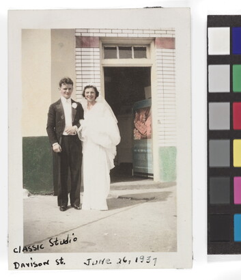 A color photograph of a young White man and woman dressed in wedding attire and standing in front of a doorway.