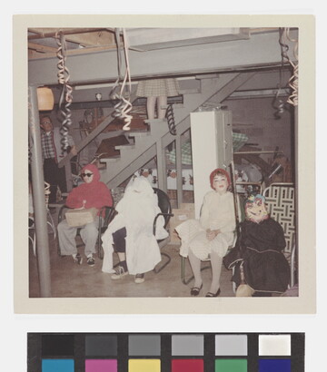 A faded color photograph of four children in costumes sitting in chairs at the foot of basement stairs.