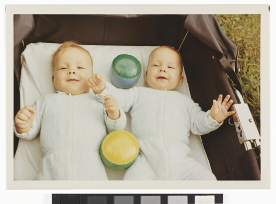 A color photograph of two White babies smiling and lying on their backs with toys in a baby carriage that is sitting in grass.